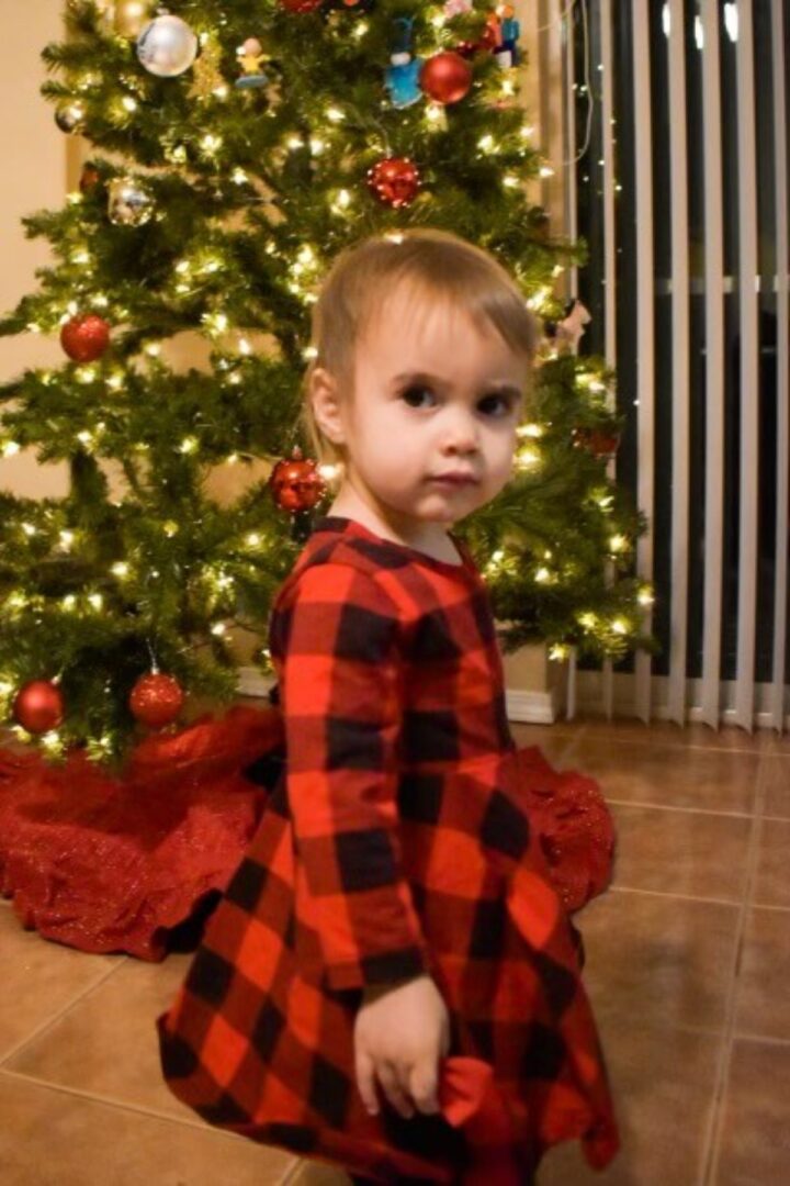 A little girl wearing a red dress posing next to a Christmas tree